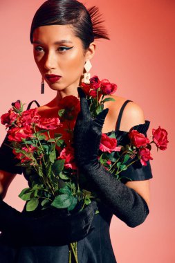 charming and trendy asian woman with bold makeup, in trendy earrings, long gloves and black cocktail dress posing with red and fresh roses on pink background, generation z, trendy spring concept clipart