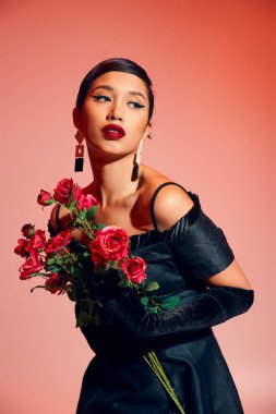 young and expressive asian woman with brunette hair, bold makeup, in black cocktail dress, long gloves and trendy earrings holding roses and looking away on pink background, stylish spring concept clipart
