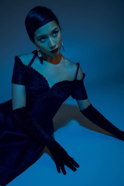 Zarif bahar kıyafetleri içinde genç Asyalı bir kadın mavi arka planda oturuyor ve diğer tarafa bakıyor. Cyan ışıklandırma efekti, moda küpeler, kokteyl elbisesi, siyah uzun eldivenler, gen z moda.