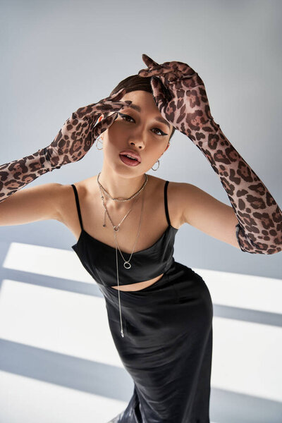 high angle view of asian woman in spring outfit, with expressive gaze looking at camera on grey background with lighting, silver accessories, black strap dress, animal print gloves, fashion shoot