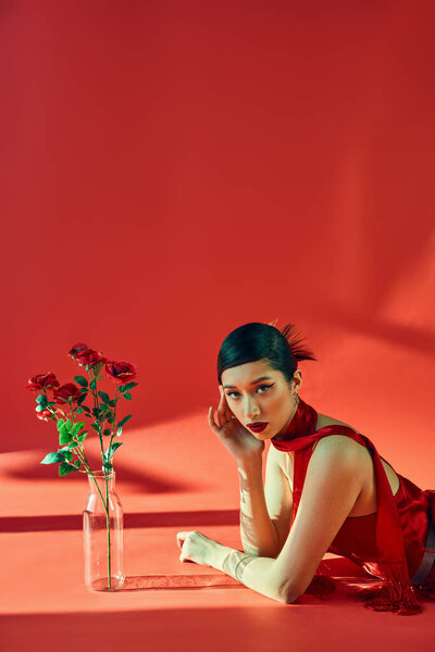 spring fashion concept, young asian woman with brunette hair and bold makeup laying in neckerchief and dress while looking at camera near fresh roses on red background with lighting