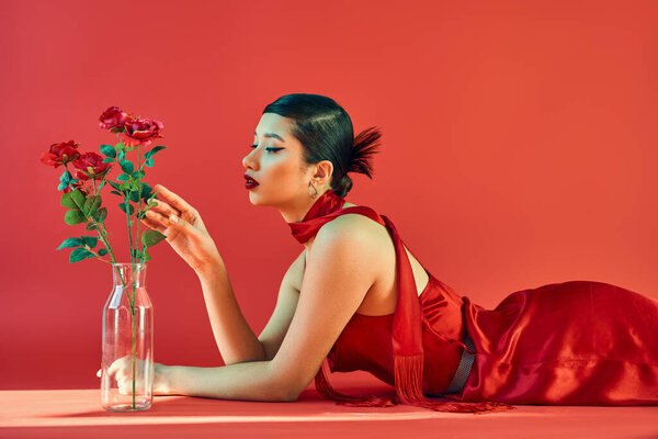 side view of charming asian woman with brunette hair and bold makeup, in elegant dress and neckerchief lying and touching roses in glass vase on red background with lighting, spring fashion concept