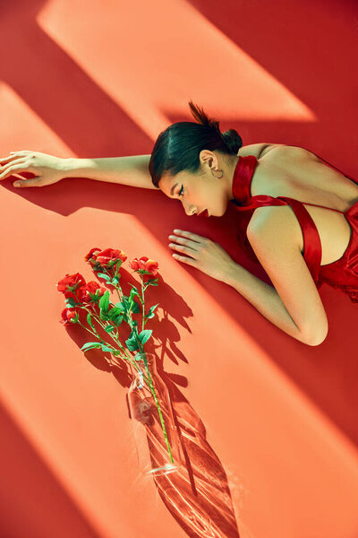 top view of expressive and young asian woman with brunette hair, in neckerchief and dress lying in lighting near glass vase with roses on red background, generation z, spring fashion photography
