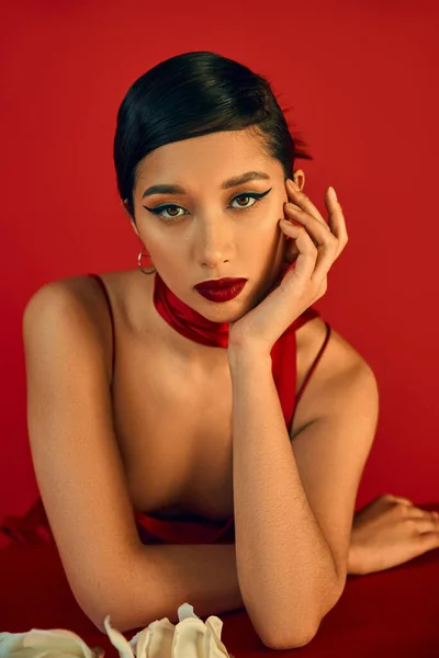 迷人而时尚的亚洲女人的肖像 穿着皮带衫和领巾 乌黑的头发和大胆的化妆 手拉手挨着脸坐着 看着红色背景的相机 春天时尚 — 图库照片