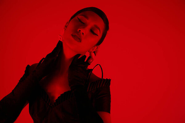 stylish and trendy asian woman with bold makeup and closed eyes posing in long black gloves and cocktail dress on vibrant background with red lighting effect, spring fashion concept