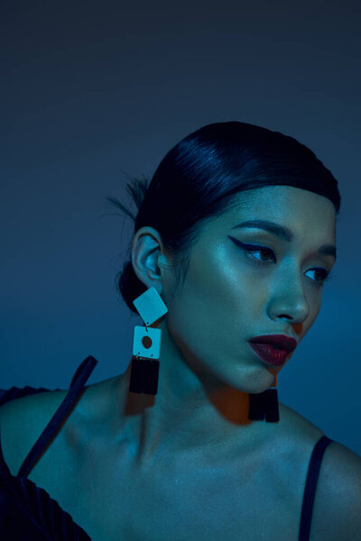 portrait of young and charming asian woman with brunette hair, bold makeup and trendy earrings looking away on blue background with cyan lighting effect, stylish spring concept