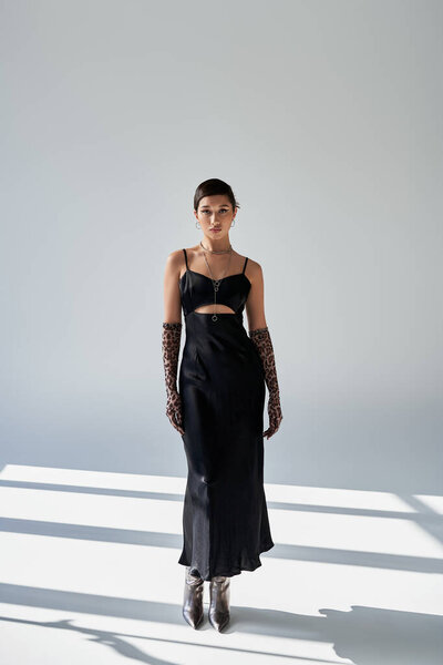 полная длина молодой азиатской женщины с брюнетками, в черном элегантном платье, перчатки для печати животных и серебряные сапоги, стоящие на сером фоне с освещением и тенями, весенняя модная фотография