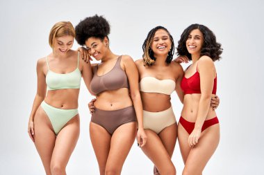 Renkli iç çamaşırlı çok ırklı kadınlar birbirlerine sarılıp kameraya bakarken gri, farklı vücut tipleri ve kendini kabullenme konsepti, çok kültürlü modeller üzerinde izole bir şekilde duruyorlar.