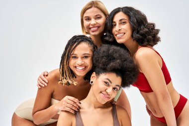 Positive multiethnic women in colorful and modern lingerie looking at camera while posing together isolated on grey, different body types and self-acceptance concept, multicultural models clipart