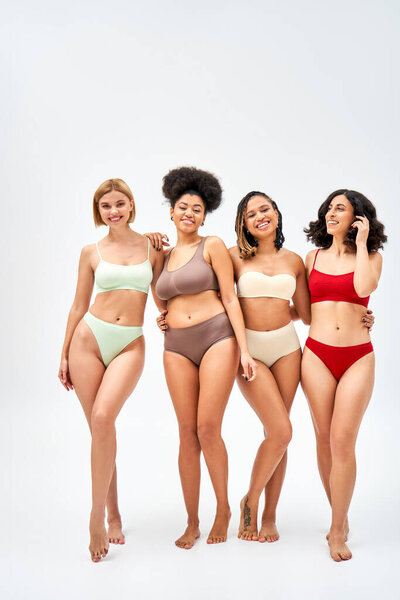 Full length of joyful multiethnic women in colorful bras and panties hugging while standing together on grey background, different body types and self-acceptance concept, multicultural models