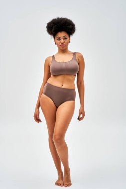 Kendinden emin ve çıplak ayaklı Afro-Amerikan kadın. Kahverengi iç çamaşırlı. Gri arka plan, kendini kabullenme ve vücut pozitifliği üzerine poz verirken kameraya bakıyor. 