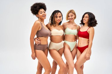Renkli iç çamaşırları içinde neşeli ve modern çok ırklı kız arkadaşlar. Gri, çok kültürlü modeller ve vücut pozitifliği konseptiyle izole edilmiş bir şekilde poz verirken birbirlerine sarılıyorlar.