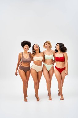 Full length of cheerful and barefoot multiethnic women in colorful lingerie hugging and walking together on grey background, multicultural models and body positivity movement concept clipart