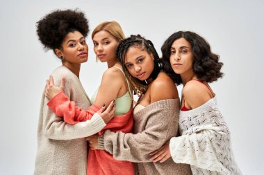 Portrait of confident and multiethnic women in knitted sweaters hugging and looking at camera isolated on grey, different body types and self-acceptance, multicultural representation clipart