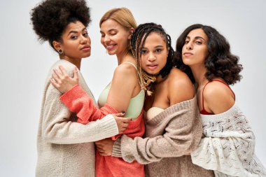 Portrait of group of multiethnic girlfriends in warm knitted jumpers hugging and standing together isolated on grey, different body types and self-acceptance, multicultural representation clipart