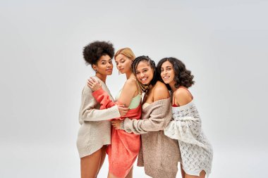 Cheerful and stylish multiethnic women in wool sweaters hugging and standing together isolated on grey, different body types and self-acceptance, multicultural representation clipart