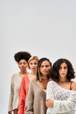Multiethnic women in warm knitted sweaters looking at camera and standing next to each other isolated on grey, different body types and self-acceptance, multicultural representation clipart