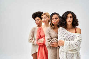 Stylish multiethnic women in trendy knitted jumpers crossing arms and looking away while standing isolated on grey, different body types and self-acceptance, multicultural representation clipart