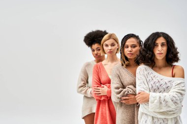 Multiethnic group of women in stylish knitted sweaters crossing arms and looking at camera isolated on grey, different body types and self-acceptance, multicultural representation, copy space clipart