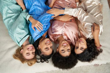 Renkli pijama giymiş neşeli çoklu etnik kız arkadaşların evde pijama partisinde, pijama partisinde, pijama partisinde, pijama partisinde ve pijama partisinde birbirlerine sarılıp kameraya bakışlarının üst görüntüsü.