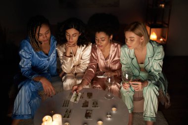 divination, african american woman in pajama holding tarot cards near multicultural girlfriends with wine and candles on table during girls night at home, bonding time in comfortable sleepwear clipart