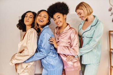 Renkli pijama giymiş mutlu çok kültürlü kız arkadaşlar kameraya bakıyor, pijama partisinde birbirlerine sarılıyorlar rahat uyku kıyafetleri ve kültürel çeşitlilikte zaman geçiriyorlar.