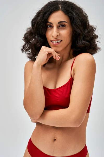 Portrait of positive brunette and middle eastern woman in modern red lingerie looking at camera while posing and standing isolated on grey, self-acceptance and body positive concept