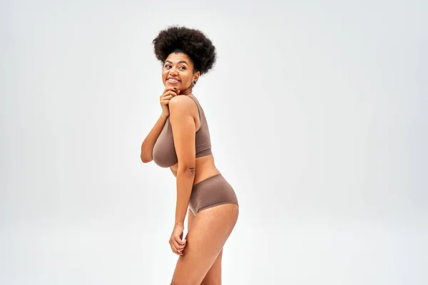 Cheerful african american model in brown lingerie touching chin and looking away while standing and posing isolated on grey, self-acceptance and body positive concept