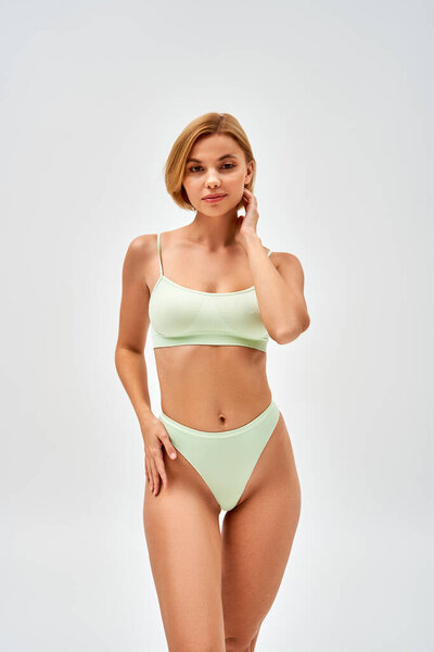 Pretty young blonde woman with natural makeup in light green lingerie touching hip while standing and looking at camera isolated on grey, self-acceptance and body positive concept 