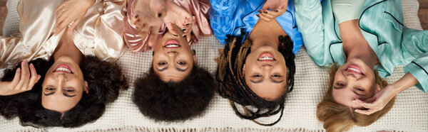 Вид сверху многоэтнических и позитивных подруг в цветной пижаме, протягивающих руки перед камерой, лежа на кровати во время пижамной вечеринки дома, проводя время в удобной сонной одежде, баннер