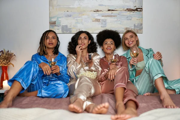 Smiling and multiethnic girlfriends in colorful pajamas holding wine and popcorn watching tv while sitting on bed during pajama party at home, bonding time in comfortable sleepwear