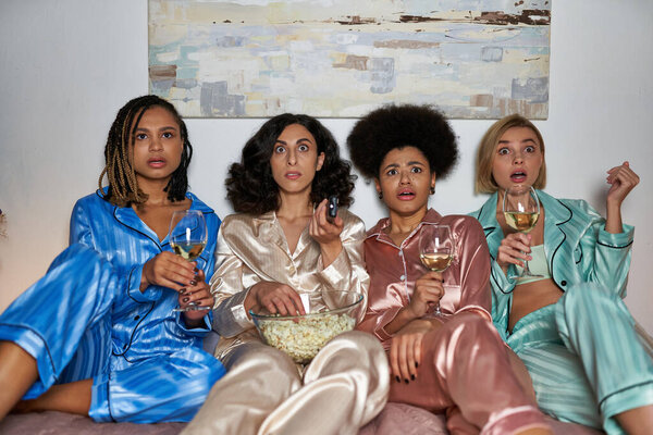 Scared multiethnic girlfriends in colorful pajama holding wine and popcorn while watching tv and sitting on bed during girls party at home, bonding time in comfortable sleepwear, slumber party