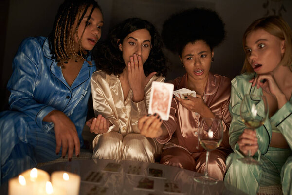 divination, african american woman in pajama holding blurred tarot card near shocked multiethnic girlfriends, wine glasses and candles during girls night at home, bonding time in sleepwear