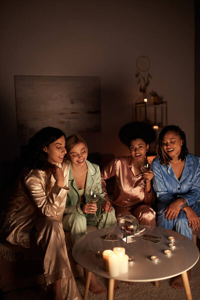 Positive multiethnic girlfriends in colorful pajama holding glasses of wine near tarot cards, crystal ball and candles during girls night at home, bonding time in comfortable sleepwear, divination 