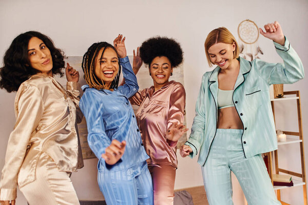 Cheerful multicultural girlfriends in colorful pajama dancing and having fun together while looking at camera during pajama party at home, slumber party, bonding time in comfortable sleepwear