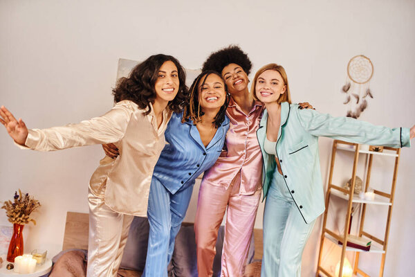 Smiling multicultural girlfriends in colorful pajama hugging and looking at each other together while standing on bed during slumber party at home, bonding time in comfortable sleepwear
