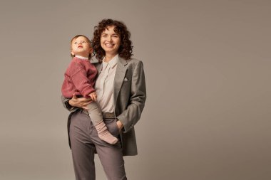 İş hayatı dengesi konsepti, takım elbiseli neşeli bir kadın kızını tutuyor ve gri arka plan, kariyer ve ailede el ele tutuşuyor sevgi dolu annelik, güçlü kadın, kendine güvenen lider. 