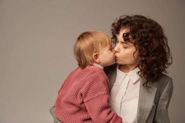 work life balance concept, curly woman in suit kissing her daughter on grey background, loving mother, balanced lifestyle, modern working woman. modern parenting, career and family  clipart