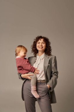 Kaliteli zaman, iş hayatı dengesi konsepti, takım elbiseli mutlu kadın kızını tutuyor ve gri arka plan, kariyer ve ailede el ele duruyor, sevgi dolu annelik, kendine güvenen lider. 