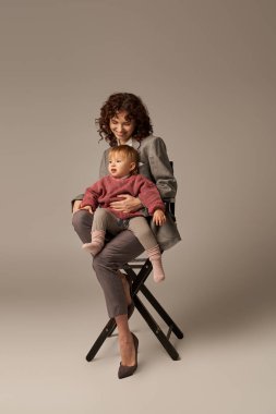 balanced lifestyle, career and family, curly woman in suit sitting on chair with daughter on grey background, professional success, motherhood, multitasking, quality time, work life balance concept  clipart
