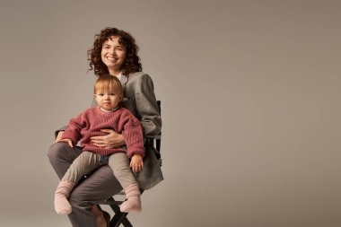 balanced lifestyle, career and family, curly businesswoman sitting on chair with child on grey background, professional success, motherhood, multitasking, quality time, work life balance concept  clipart