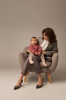 Modern çalışan ebeveyn, dengeleyici iş ve hayat konsepti, takım elbiseli kıvırcık kadın bebek kızıyla koltukta oturuyor, gri arka plan, mutlu anne ve çocuk, çoklu görev, kaliteli zaman 