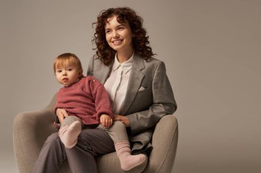 modern working parent, curly woman in suit sitting in armchair with toddler daughter, grey background, happy mother and child, multitasking, quality family time, balancing work and life concept  clipart
