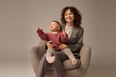 Modern çalışan ebeveyn, dengeleyici iş ve hayat konsepti, takım elbiseli mutlu kadın yeni yürümeye başlayan kızıyla koltukta oturuyor, gri arka plan, anne ve çocuk, annelik, kaliteli zaman 