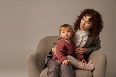 balancing work and life concept, curly woman in suit sitting in armchair with toddler daughter, grey background, multitasking, quality family time, mother and child, modern working parent  clipart