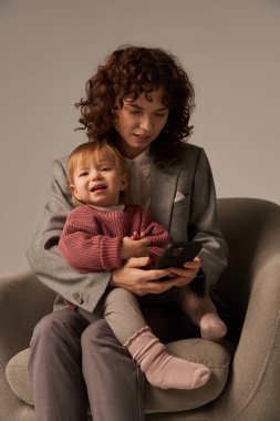 Modern çalışan anne, çoklu görev, akıllı telefon, dengeleyici iş ve hayat konsepti, takım elbiseli kadın bebek bebekle koltukta oturuyor, gri arka plan, ebeveyn ve çocuk  