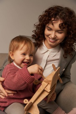 Başarılı bir anne, iş ve hayat kavramını dengelemek, takım elbiseli mutlu iş kadını koltuğunda oturan ve küçük kızıyla oynayan, tahta çift kanatlı, gri arka planlı, çocukla meşgul olan 