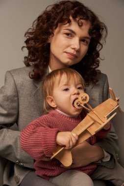 Modern çalışan anne, iş ve hayat kavramını dengelemek, takım elbiseli iş kadını, küçük kızıyla koltukta oturuyor, tahta çift kanatlı uçakla oynuyor, gri arka plan, annelik. 