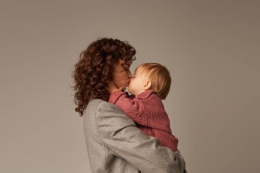 working mother, parenting and career, curly businesswoman holding in arms and kissing her toddler daughter on grey background, work life harmony concept, loving motherhood, quality time, side view clipart