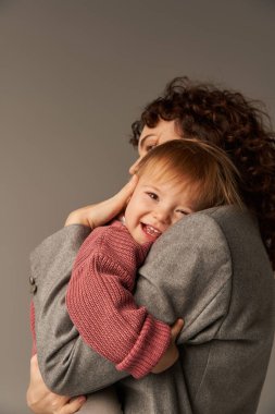 Çalışan anne, ebeveynlik ve kariyer, kıvırcık iş kadını, gri arka planda mutlu kızı kucaklayan, iş hayatı uyum konsepti, sevgi dolu annelik, kaliteli aile zamanı 